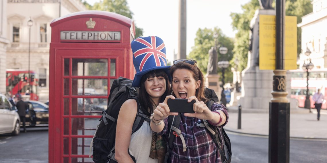 Two female tourists, one wearing a top hat featuring a union jack print pose for a selfie in front of atradition red public phone box (Photo: Getty)