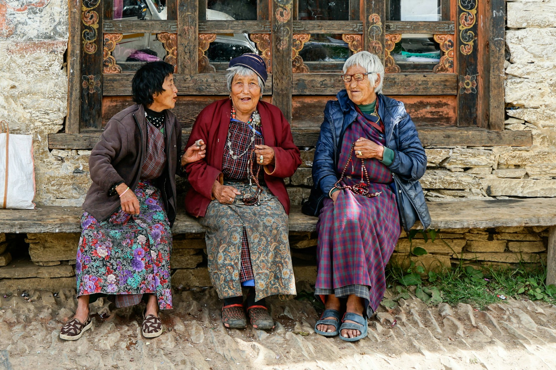 Three women sitting in front of a traditional house discussing and laughing, Mongar, Bhutan