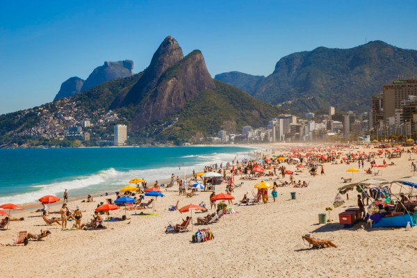 People lounging in speedos and bikinis on the beach, lying on towels or under umbrellas as the blue water crashes gently on the sand. A city and rocky mountains tower in the background under a clear blue sky.