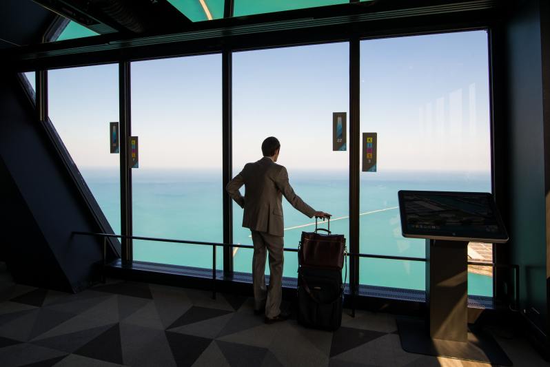 Man standing with a suitcase facing the ocean looking out windows